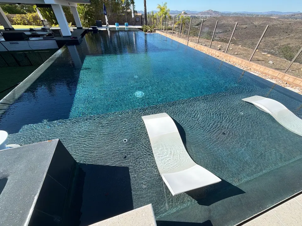 Sparkling blue water fills a dark-finished pool with a sun shelf, white loungers, and mountain views, after pool service in San Diego, CA