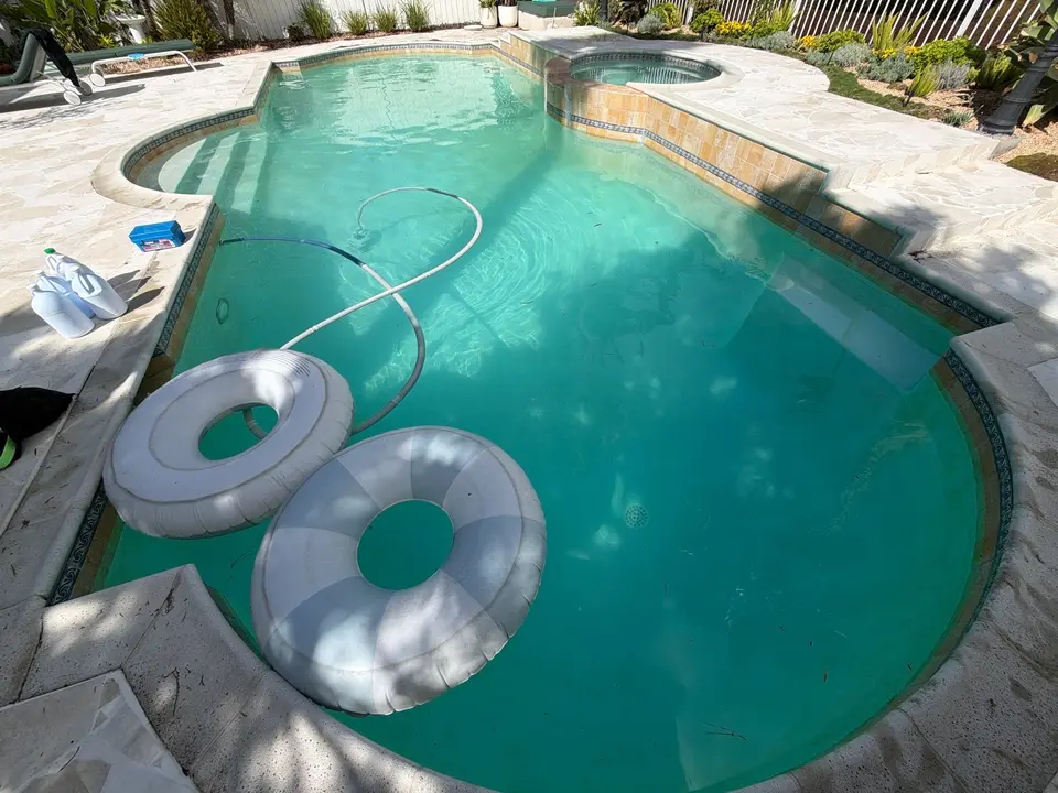 Clear turquoise water fills a curved pool with entry steps, a raised spa, stone decking, and floating rings, after pool service in San Diego, CA