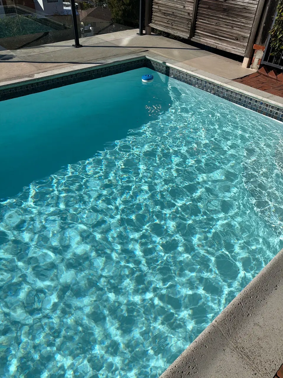 Sparkling turquoise pool water shows sunlit ripples beneath patterned waterline tile, concrete coping, and glass fencing, after pool service in San Diego, CA