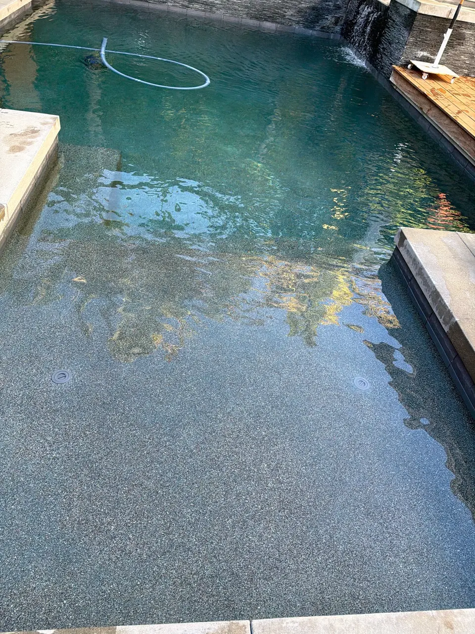 Clear teal water reveals a pebble finish and shallow shelf beneath a stone waterfall wall, after pool service in San Diego, CA