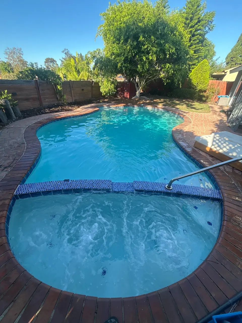 Clear turquoise water fills a freeform pool and bubbling attached spa, bordered by brick decking and mature trees, after pool service in San Diego, CA