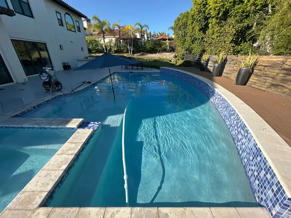 Clear blue water fills a curved pool and attached spa with mosaic tile, submerged steps, and surrounding greenery, after pool service in San Diego, CA