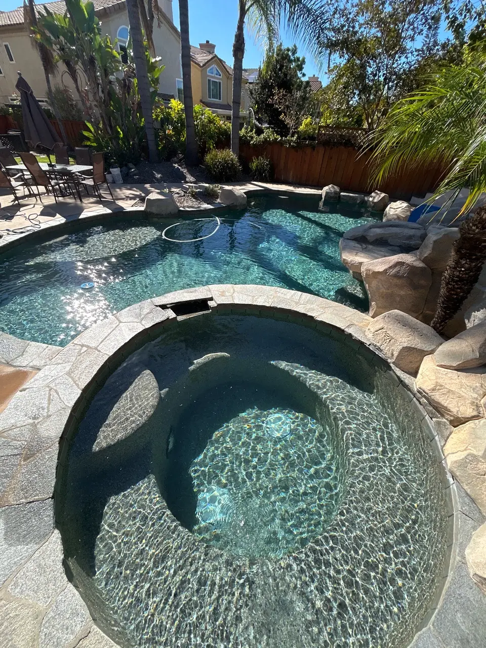 Sparkling turquoise pool and circular spa feature stone coping, rock work, a slide, and tropical landscaping, after pool service in San Diego, CA