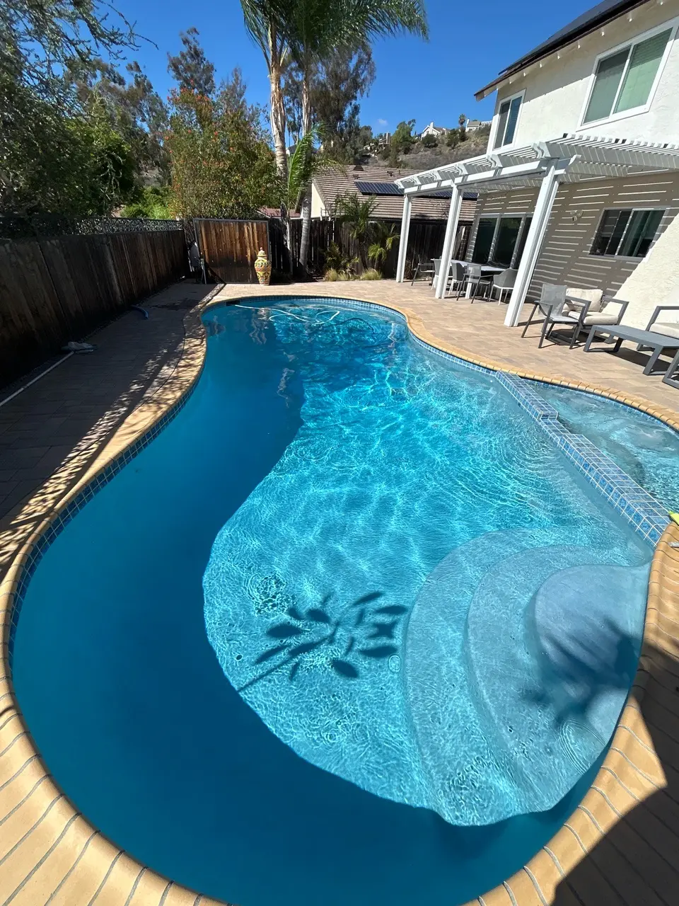 Sparkling blue water fills a freeform pool with curved entry steps, tiled seating, brick coping, and surrounding palms, after pool service in San Diego, CA