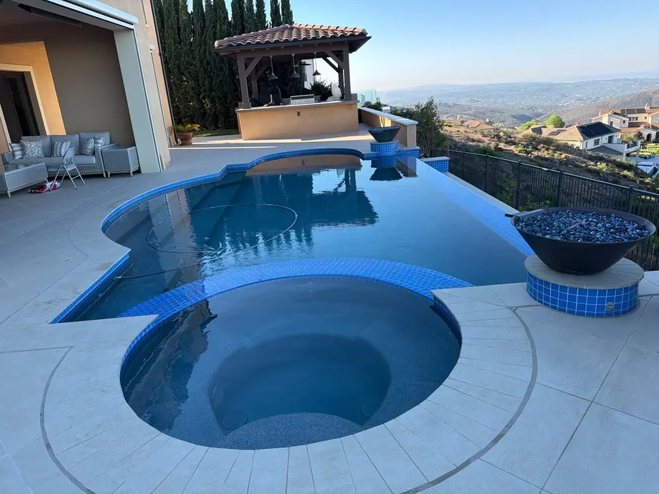 Clear blue pool and round spa feature blue tile, an infinity edge, and decorative bowls overlooking hills, after pool service in San Marcos, CA