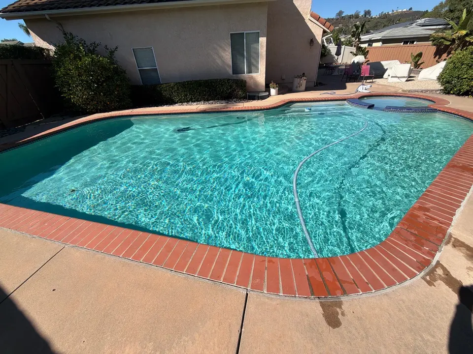 Sparkling turquoise pool with attached raised spa, brick coping, underwater steps, and a visible cleaning hose, after pool service in San Marcos, CA