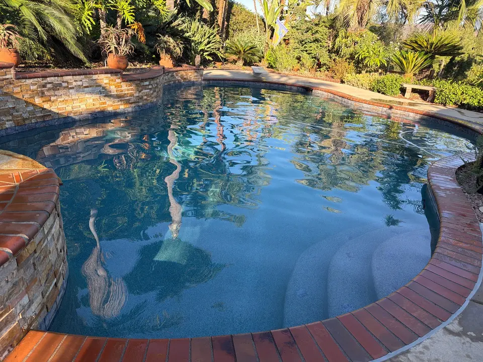Sparkling blue pool with submerged steps, brick coping, stacked-stone walls, and lush tropical landscaping, after pool service in San Marcos, CA