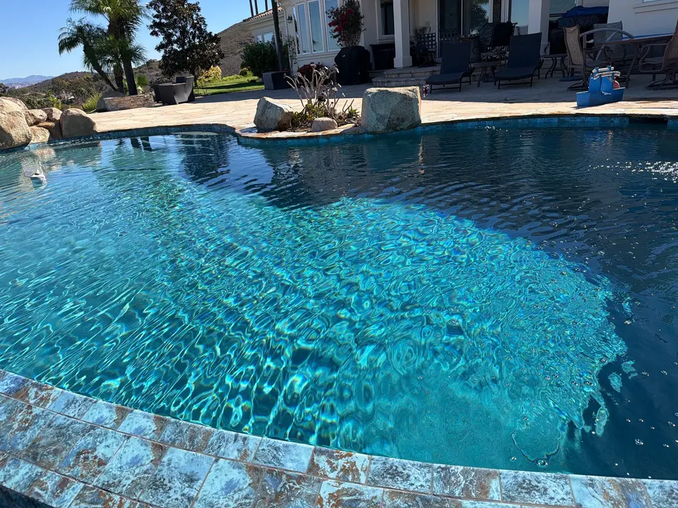 Sparkling blue water fills a freeform pool bordered by decorative tile, natural rocks, and palm landscaping, after pool service in San Marcos, CA