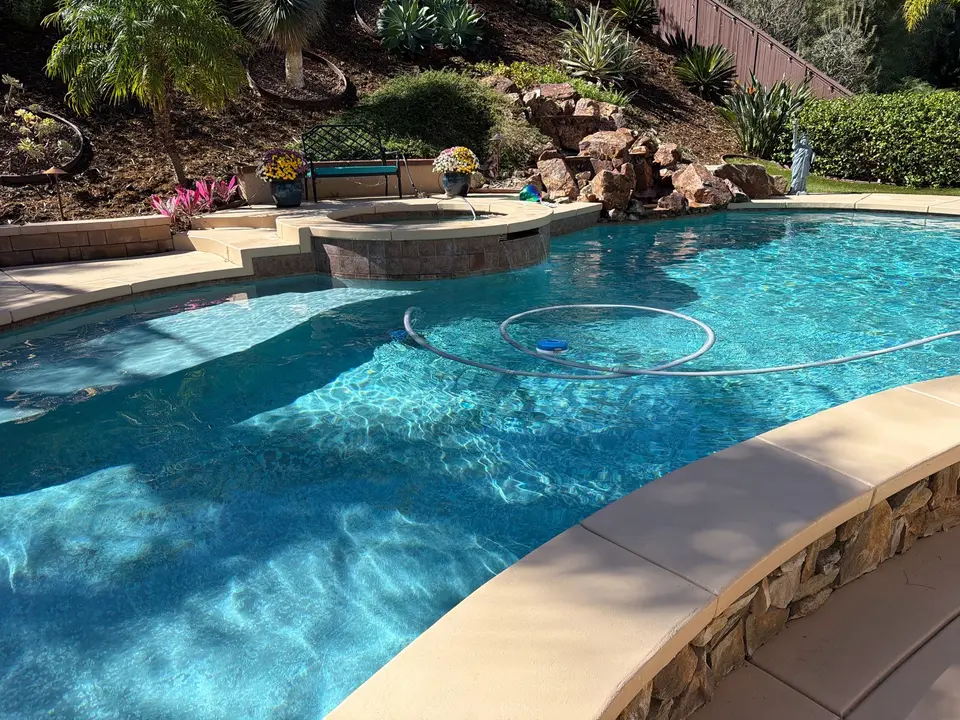 Sparkling blue pool water surrounds submerged steps beneath a raised spa, rock waterfall, and tropical landscaping, after pool service in San Marcos, CA