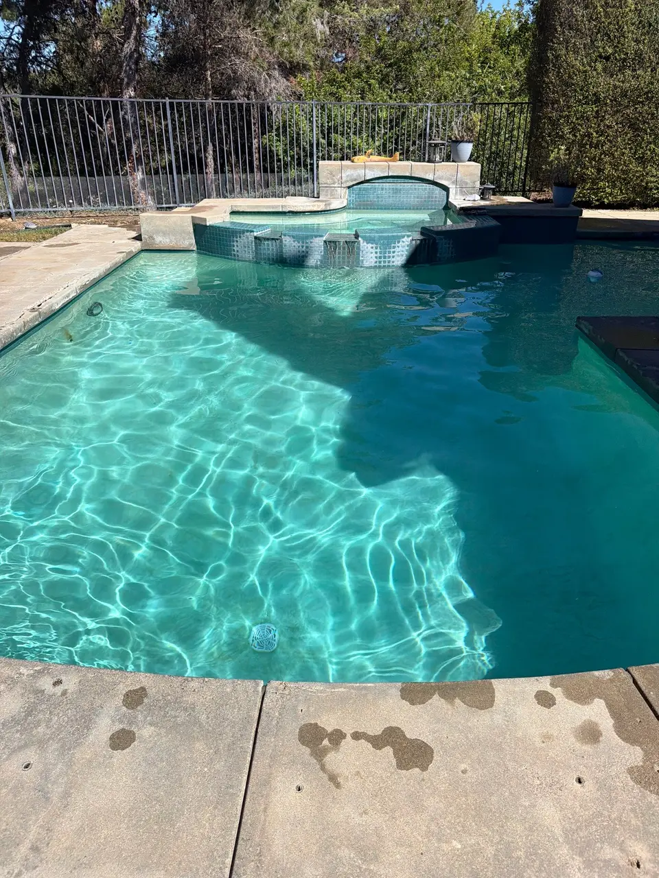 Sparkling turquoise pool water surrounds a raised tiled spa with a spillway, bordered by concrete decking and greenery, after pool service in San Marcos, CA