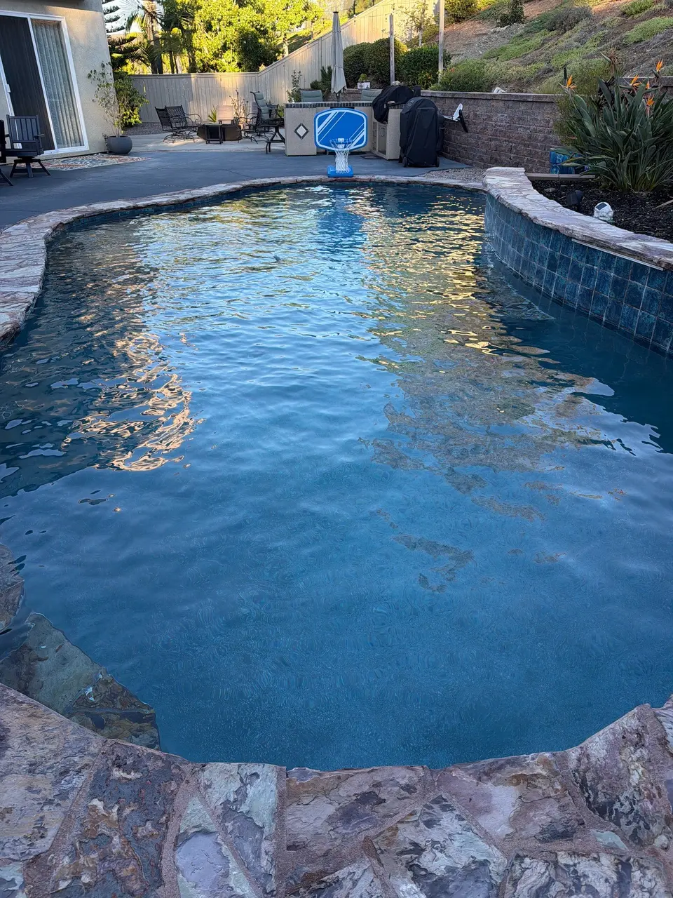 Clear blue water fills a freeform pool with stone coping, a raised tiled wall, and basketball hoop, after pool service in San Marcos, CA