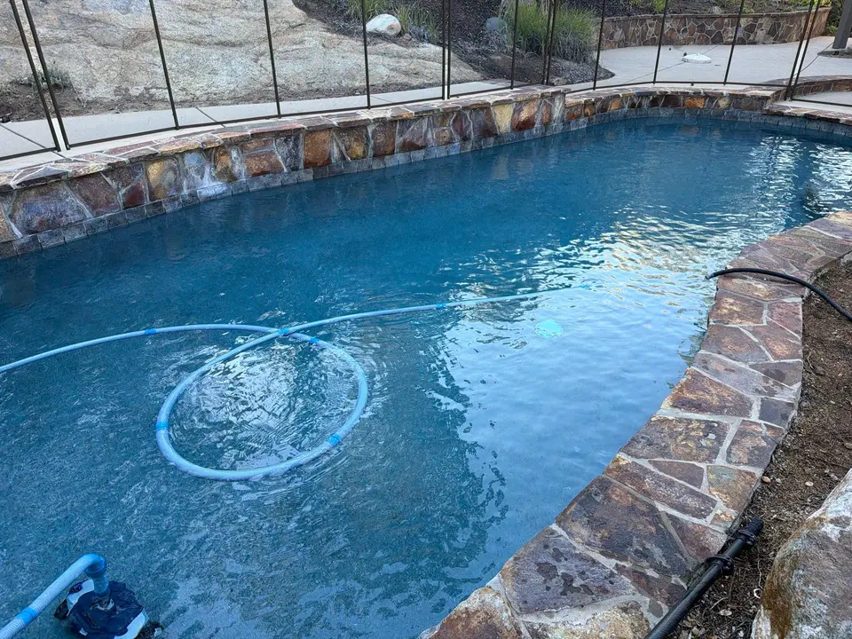 Clear blue water fills a curved pool with stone coping, a raised stone wall, and a vacuum hose, after pool service in San Marcos, CA