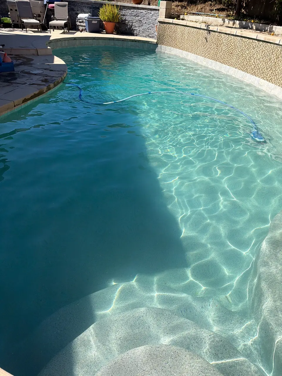 Clear turquoise water reveals curved entry steps and a light finish beside a mosaic tile wall, after pool service in San Marcos, CA