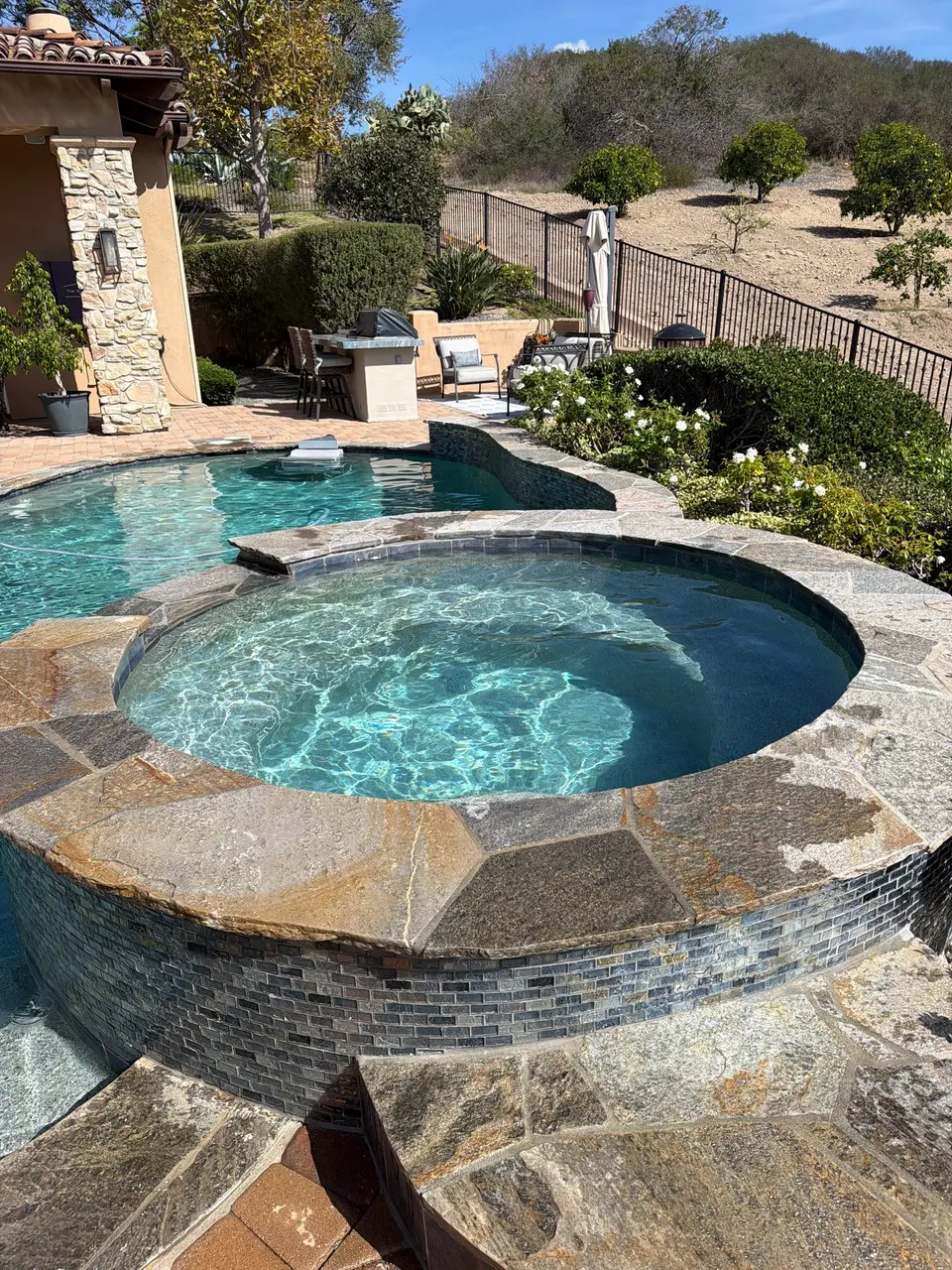 Sparkling turquoise water fills a raised circular spa with stone coping and mosaic tile beside a landscaped pool, after pool service in Santee, CA