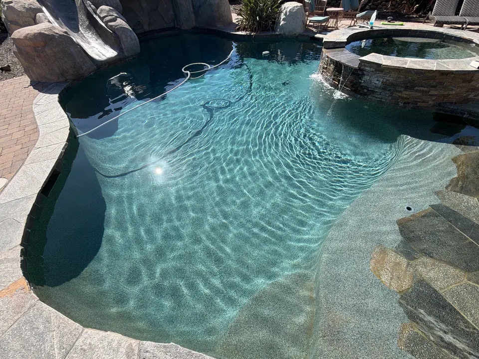 Sparkling turquoise pool with a pebble finish, rock slide, submerged steps, and raised stone spa with spillover, after pool service in Scripps Ranch, CA