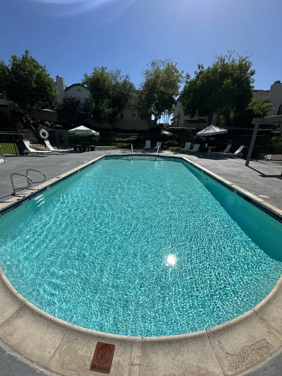 Sparkling turquoise pool with a rounded end, blue waterline tile, metal handrails, and surrounding lounge chairs and umbrellas, after pool service in Spring Valley, CA