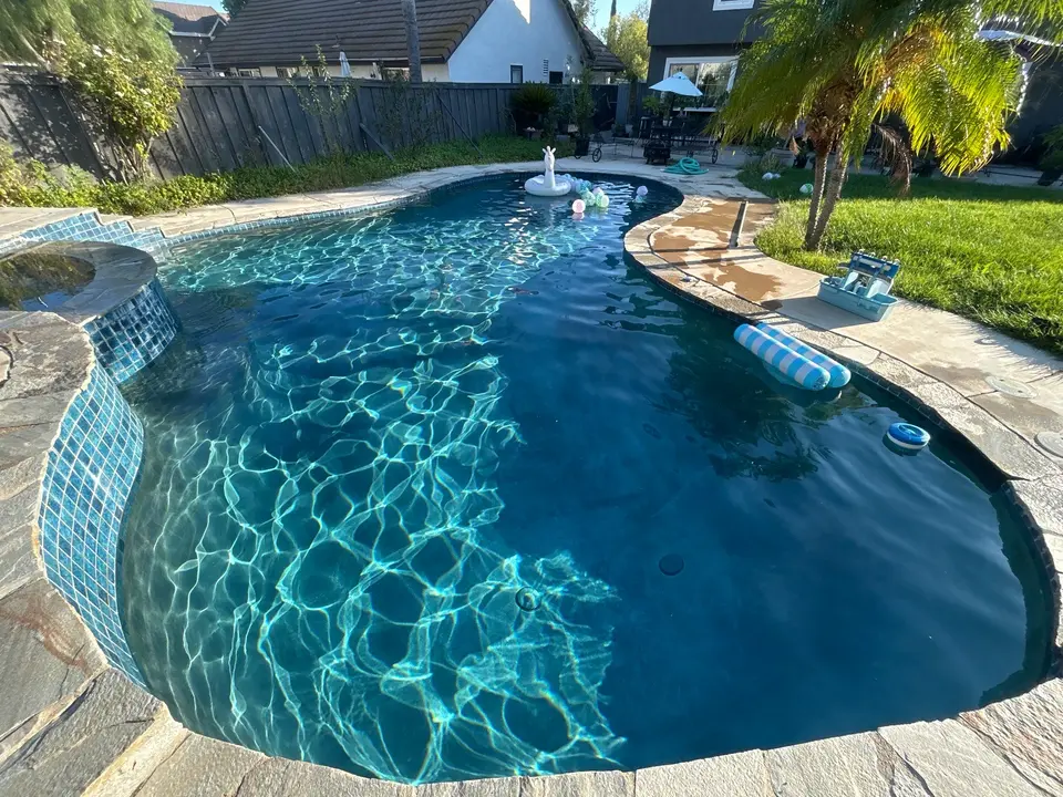 Sparkling blue water fills a freeform pool beside a raised tiled spa, stone coping, and palm trees, after pool service in Temecula, CA