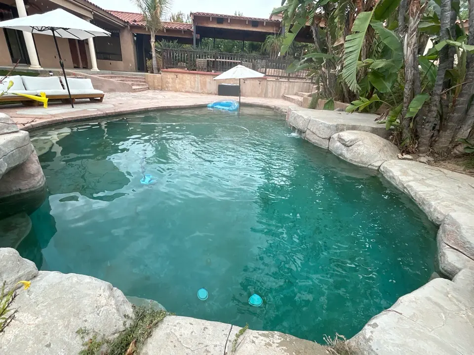 Clear turquoise water fills a freeform pool bordered by rock coping, a small waterfall, and tropical landscaping, after pool service in Temecula, CA
