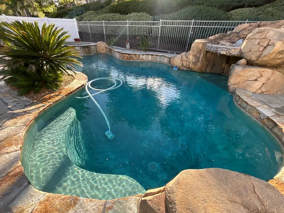 Clear blue water fills a freeform pool with curved steps, stone coping, rock waterfall, and tropical landscaping, after pool service in Temecula, CA