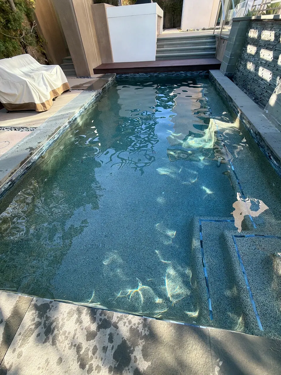 Clear blue water fills a rectangular pebble-finish pool with submerged steps beside a stacked-stone wall, after pool service in Torrance, CA