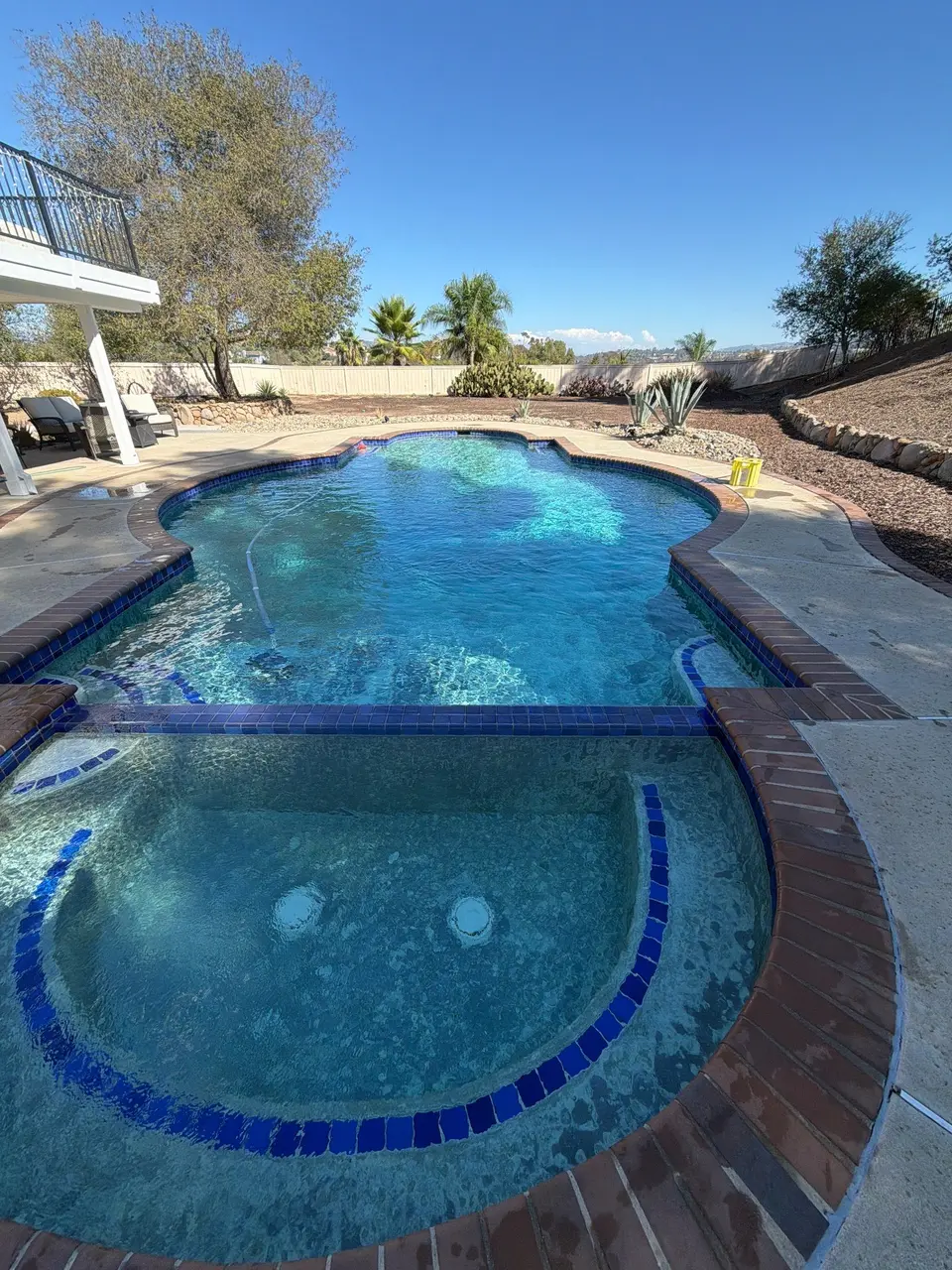 Sparkling blue water fills a curved pool and attached spa with blue tile, brick coping, and desert landscaping, after pool service in Vista, CA