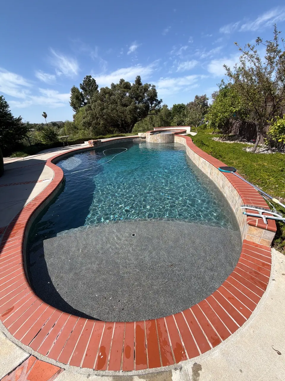 Sparkling blue water fills a curved pool with a shallow entry, brick coping, and raised circular spa, after pool service in Vista, CA