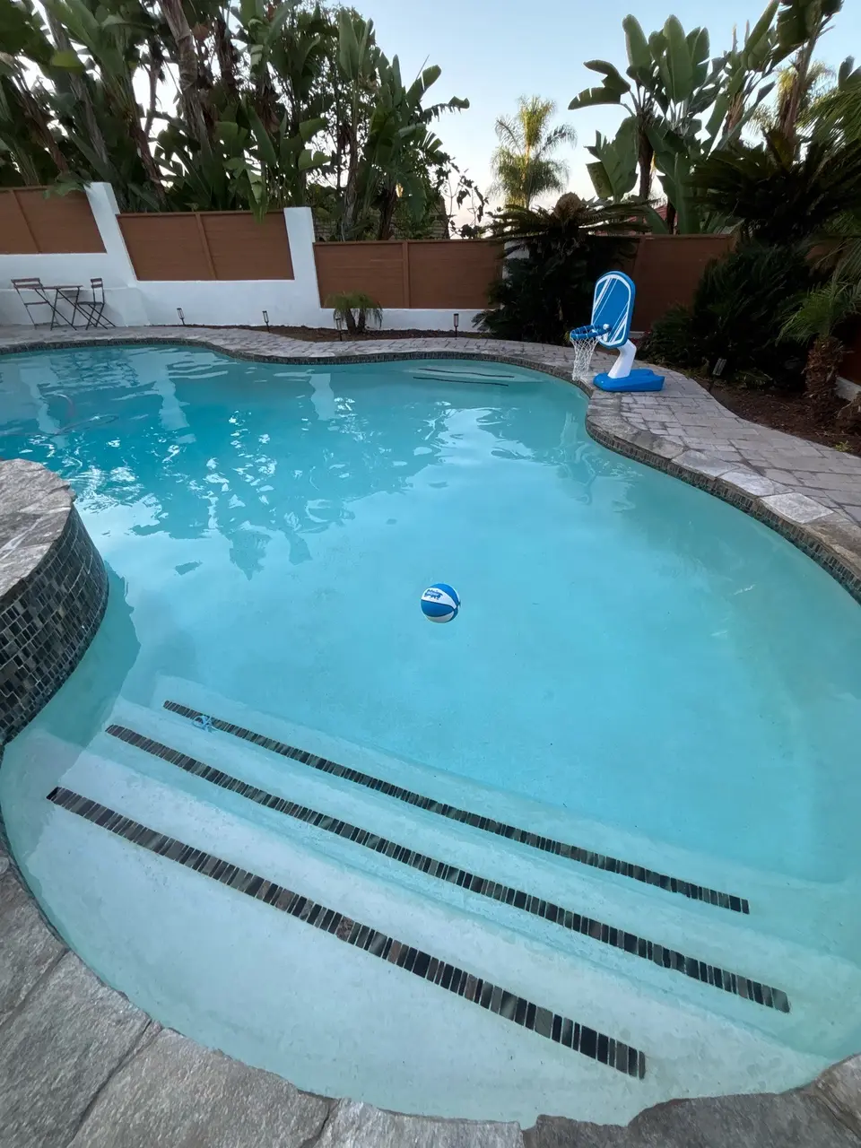 Clear turquoise pool with wide tiled entry steps, a basketball hoop, and tropical landscaping, after pool service in Vista, CA