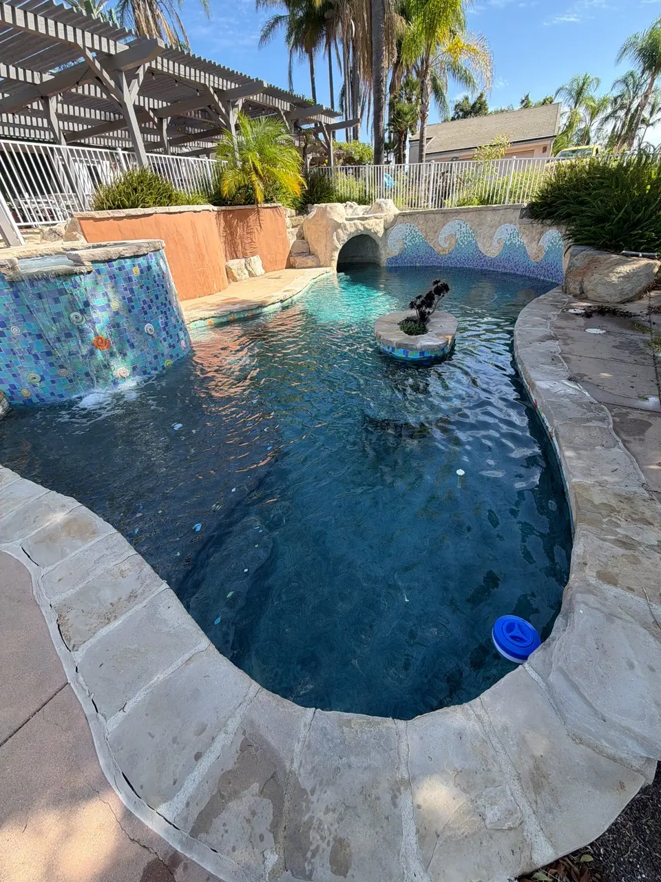Clear blue pool with colorful mosaic tile, a rock grotto, waterfall, central sculpture, and surrounding palms, after pool service in Vista, CA