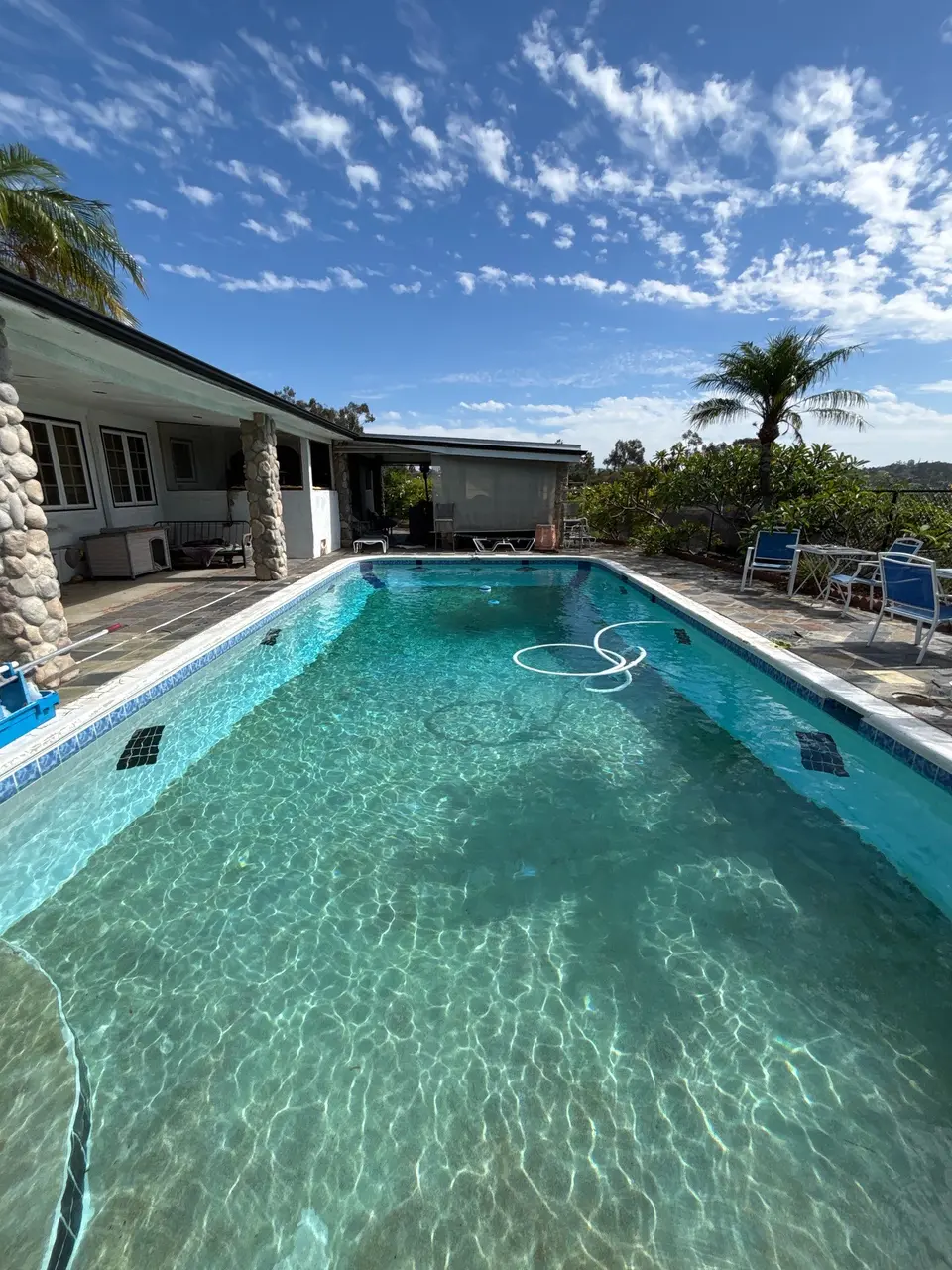 Sparkling turquoise water fills a rectangular pool with blue tile, a vacuum hose, stone patio, and palms, after pool service in Vista, CA