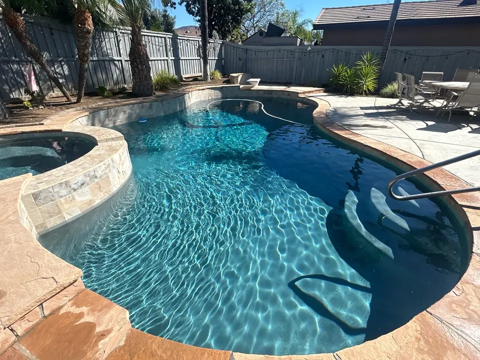 Sparkling blue water fills a curved pool with entry steps, a raised tiled spa, and surrounding palms, after pool service in Winchester, CA